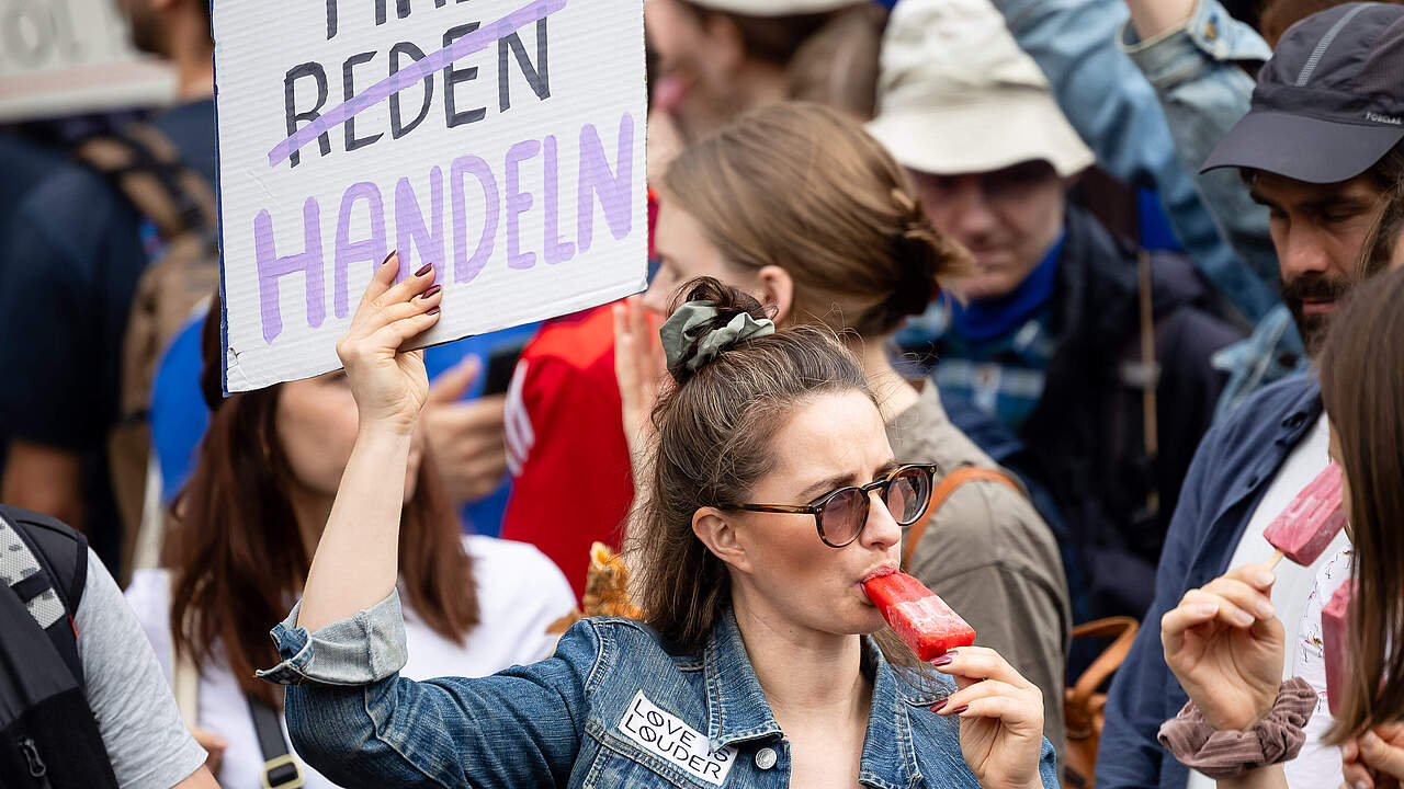 Eine junge Frau in einer Menschenmenge hält ein Schild auf dem steht: Wir müssen man handeln. Sie trägt eine Jeansjacke und isst ein Eis. 