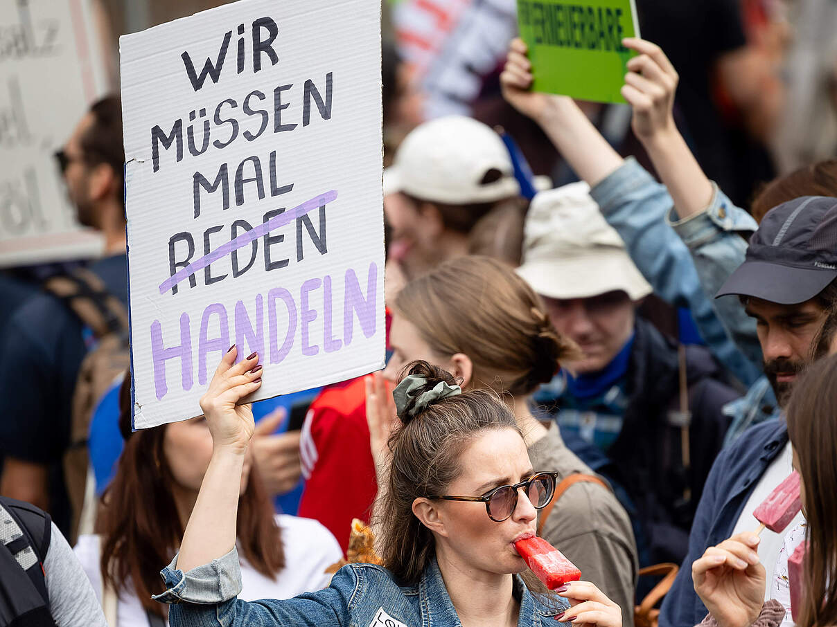 Eine junge Frau in einer Menschenmenge hält ein Schild auf dem steht: Wir müssen man handeln. Sie trägt eine Jeansjacke und isst ein Eis. 